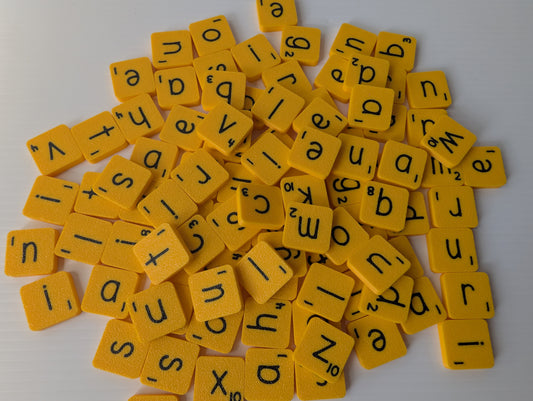 Tas de tuiles alphabétiques jaunes vues de près, lettres minuscules avec valeur en points inscrite dans un coin de chaque tuile.